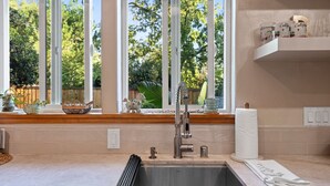 A kitchen with a stainless steel sink, a window with a view of trees, and a shelf with kitchen items.