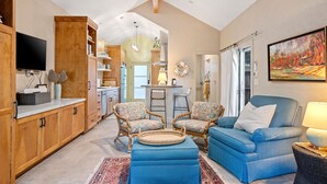 A spacious living room with a blue sofa, two patterned armchairs, and a coffee table. A television is mounted on the wall, and there is a kitchen area visible in the background.