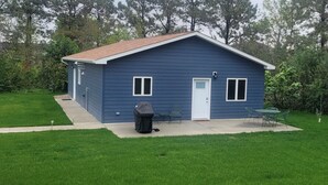 Une petite maison bleue d’un seul étage, avec une porte et des fenêtres blanches, un barbecue couvert et une terrasse avec des chaises.