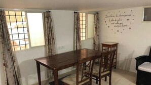 A dining area with a wooden table and chairs, two windows with curtains, and a wall with a decorative quote. - Z2 - Maison de ZEN 2, For Serenity, Bliss & Relaxation (Murugeshpalya)