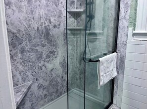 A glass-enclosed shower with a marble wall, a towel rack, and a white tiled wall. - Stylish Bungalow Near Fort Benning (Columbus)