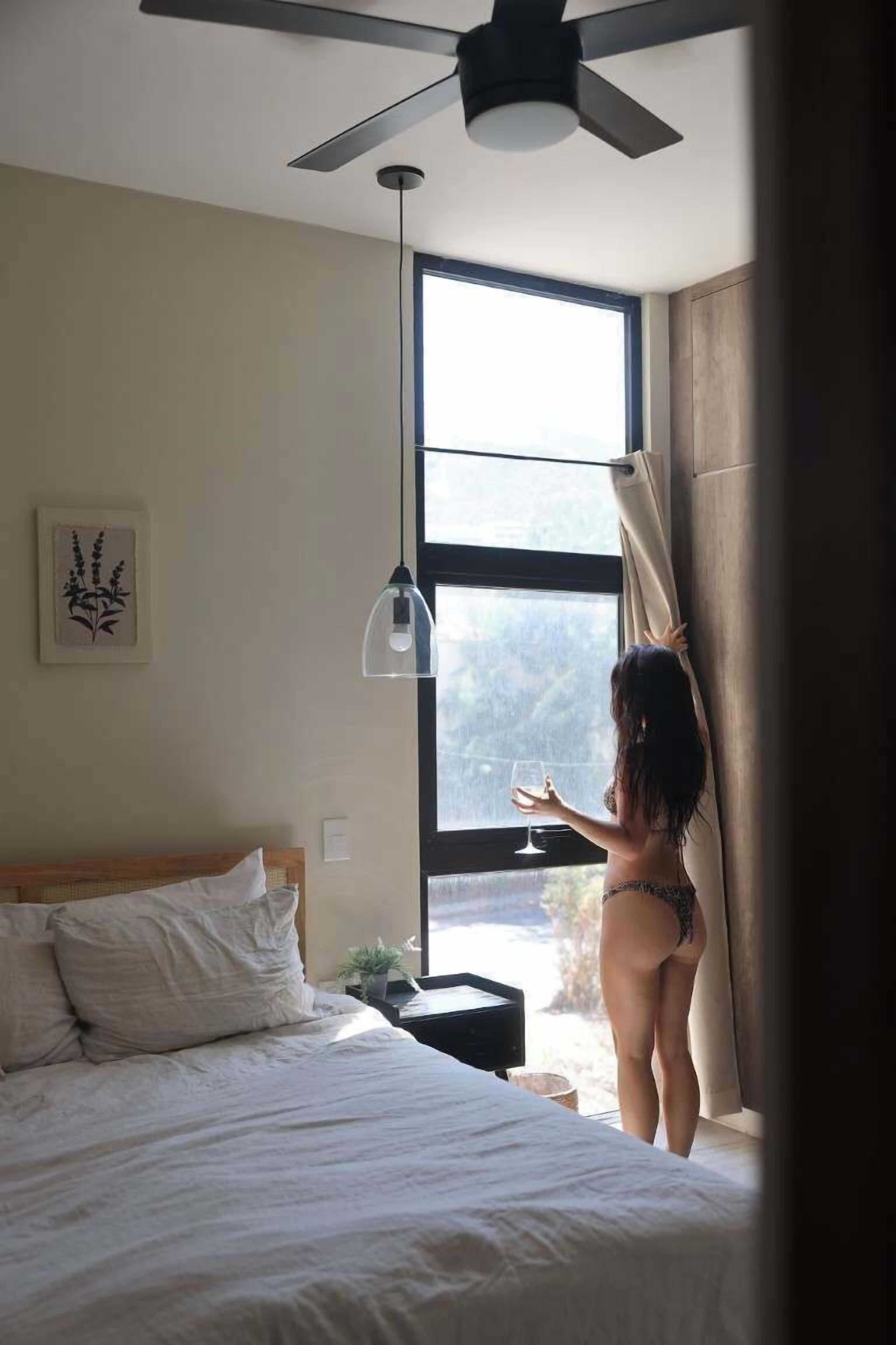 Ein Schlafzimmer mit Bett, Deckenventilator, Fenster mit Vorhängen und Nachttisch mit Lampe.