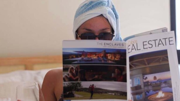 A person reading a magazine titled "REAL ESTATE" in a room with a bed, a hanging light fixture, and a framed artwork on the wall. - Ocean's Point by Baja Toms (Cabo San Lucas)