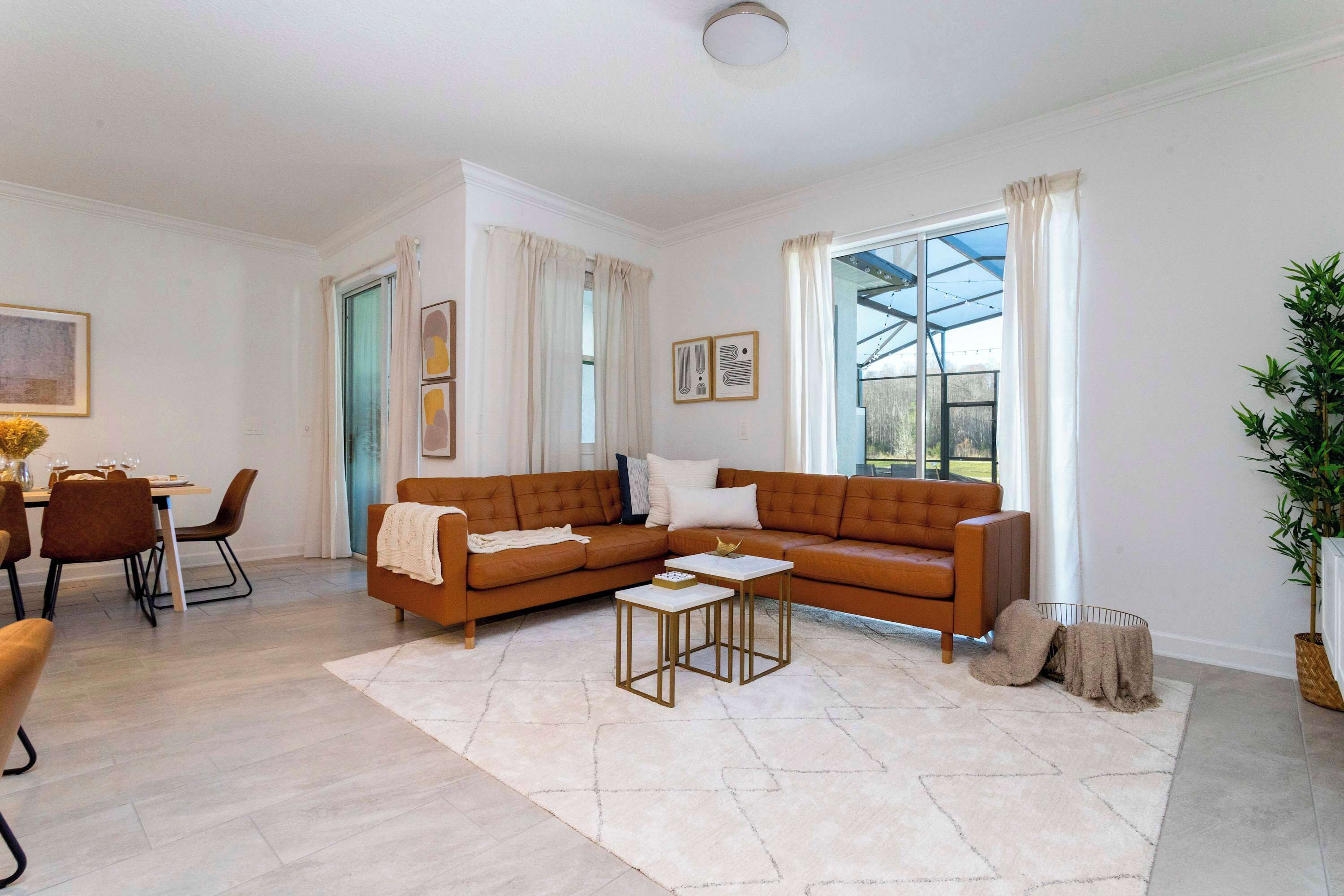 A modern living room with a brown sofa, two side tables, a dining area with a table and chairs, and a large window with curtains.