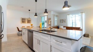 A modern kitchen with white cabinetry, a central island, pendant lighting, and a view to a living area with a sofa and curtains.