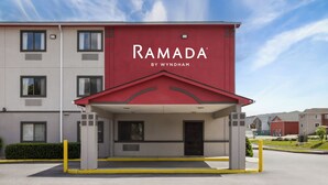Eingang eines Ramada Hotels mit roter Fassade, einem überdachten Eingangsbereich und einem Parkplatz.
