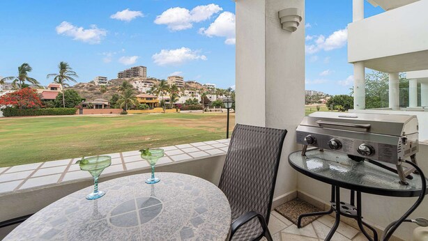 Ein Balkon mit Grill, Tisch, zwei Stühlen und Blick auf eine grüne Landschaft mit Gebäuden im Hintergrund.