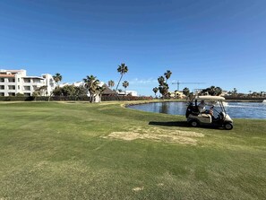Un campo de golf con un cuerpo de agua, palmeras y edificios al fondo.