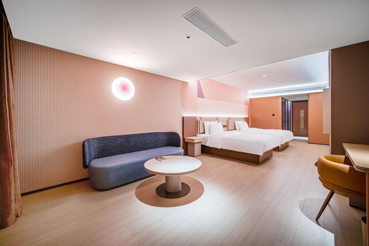 Quarto de hotel moderno com sofá azul, mesa redonda, cama com roupa de cama branca e parede de destaque em madeira.