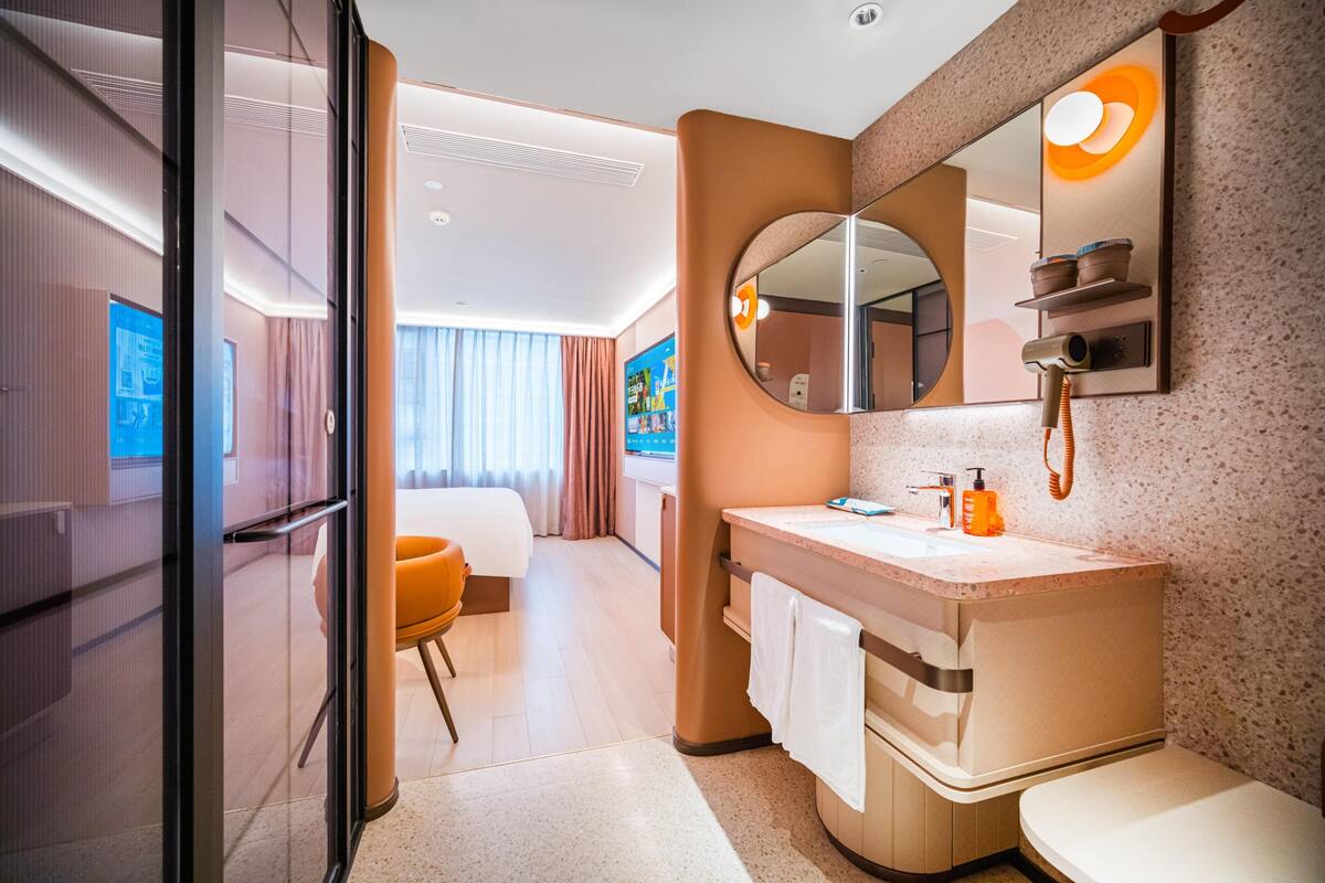 Ein modernes Hotelbad mit Waschbecken, Spiegel und Toilette. Es gibt einen Duschbereich mit Duschbrause und Handtuchhalter. Der Raum verfügt über einen großen Spiegel und ein Fenster mit Vorhängen.