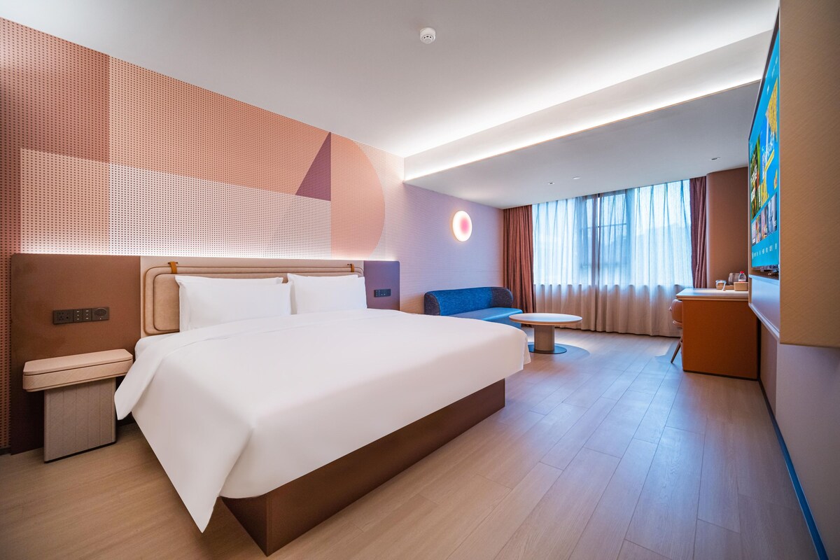 A modern hotel room with a large bed, a desk, a small sofa, and a view of the city through large windows.