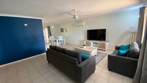 Un salon moderne avec un canapé, une table basse et un téléviseur. On aperçoit à l’arrière-plan un coin repas avec des chaises et une table.