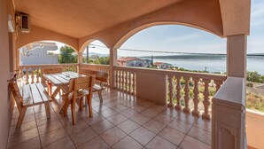 Ein überdachter Außenbereich mit Tisch und Stühlen, einem Geländer und Blick auf ein Gewässer sowie Gebäude in der Ferne.