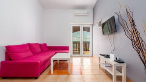 Ein modernes Wohnzimmer mit einem rosafarbenen Sofa, einem weißen Couchtisch, einem flachbildfernseher an der Wand und einem kleinen weißen Beistelltisch mit elektronischen Geräten.