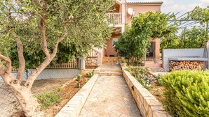 Un sendero de piedra conduce a una casa con balcón, rodeada de diversas plantas y una cerca.