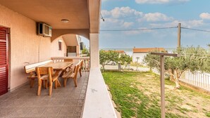 Un balcón con mesa y sillas de madera, vista al mar, y un edificio con puerta roja al fondo.