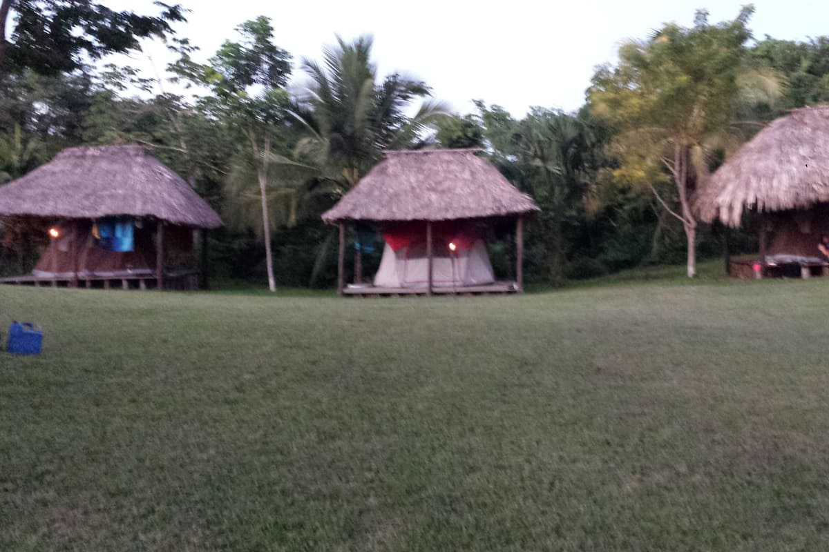 Three thatched-roof huts with open sides, set in a grassy area with trees in the background.