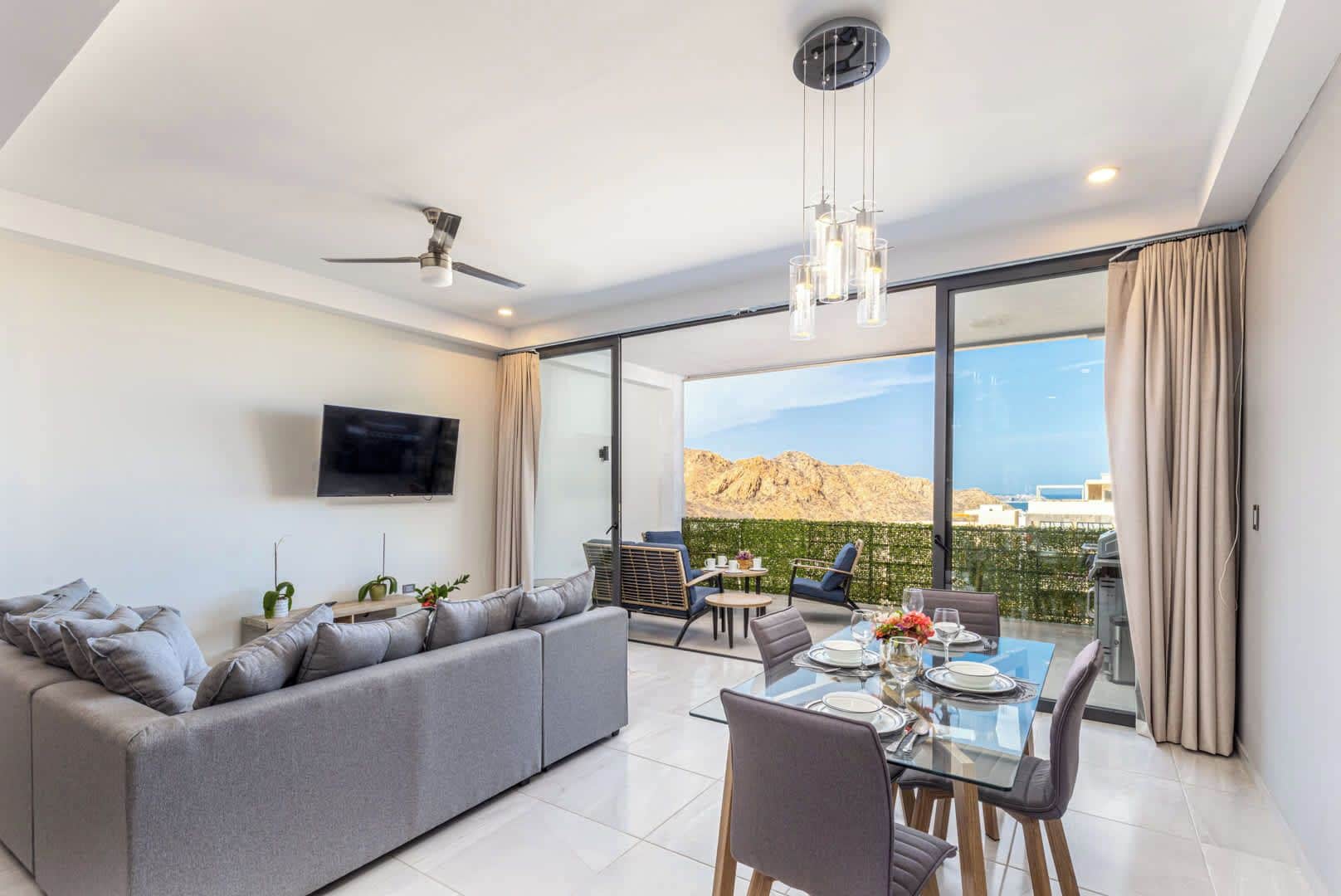 Uma sala de estar moderna com sofá, mesa de jantar e cadeiras, TV na parede e varanda com vista para as montanhas.