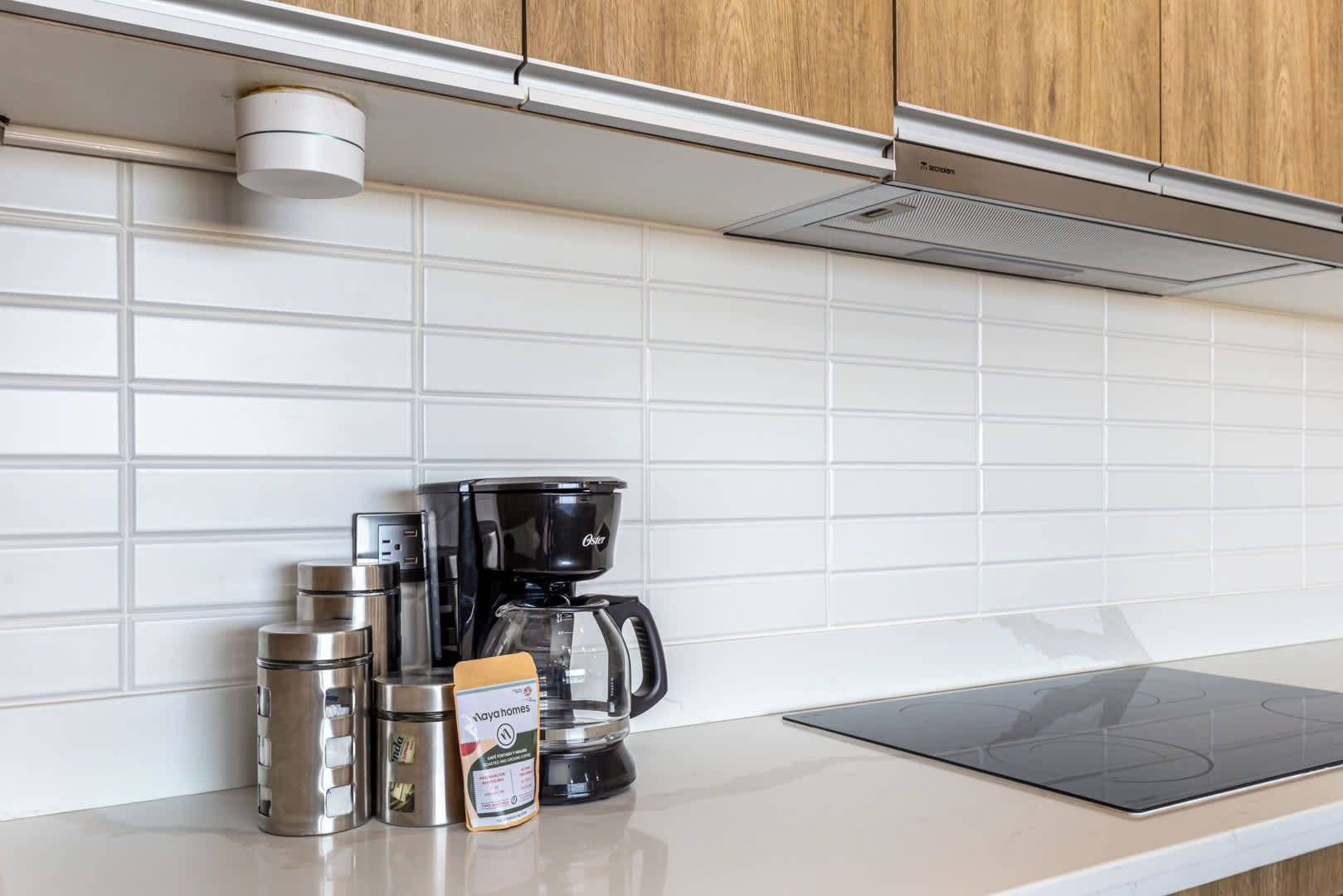 A kitchen with white tiles, a coffee maker, a kettle, a thermos, and a box of coffee pods.