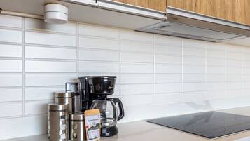 A kitchen with white tiles, a coffee maker, a kettle, a thermos, and a box of coffee pods.