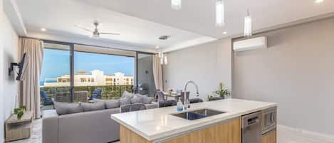 A modern kitchen with a central island, a dining area with a sofa, and a view of buildings through large windows.