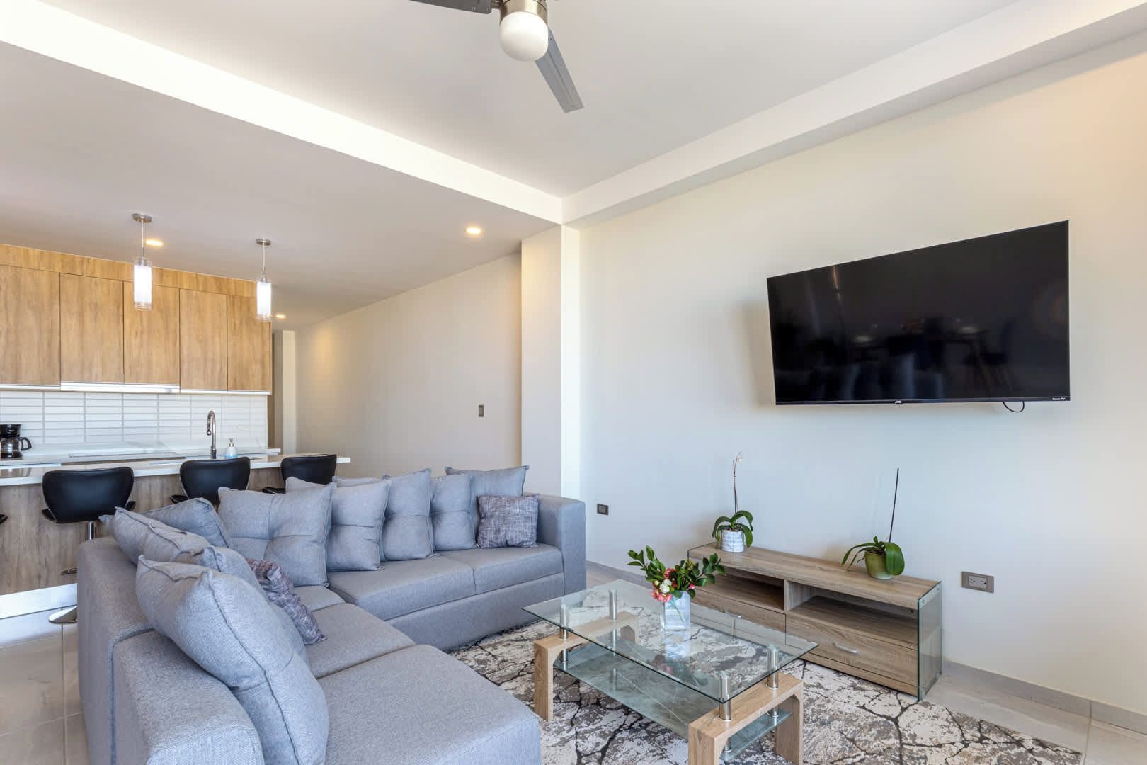 Een moderne woonkamer met een hoekbank, een glazen salontafel, een flatscreen televisie aan de muur en een keuken in de achtergrond.