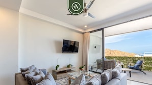 Un salon moderne avec un canapé gris, un téléviseur à écran plat et une table basse en verre. Il y a un balcon avec des sièges d’extérieur et une vue sur le paysage.