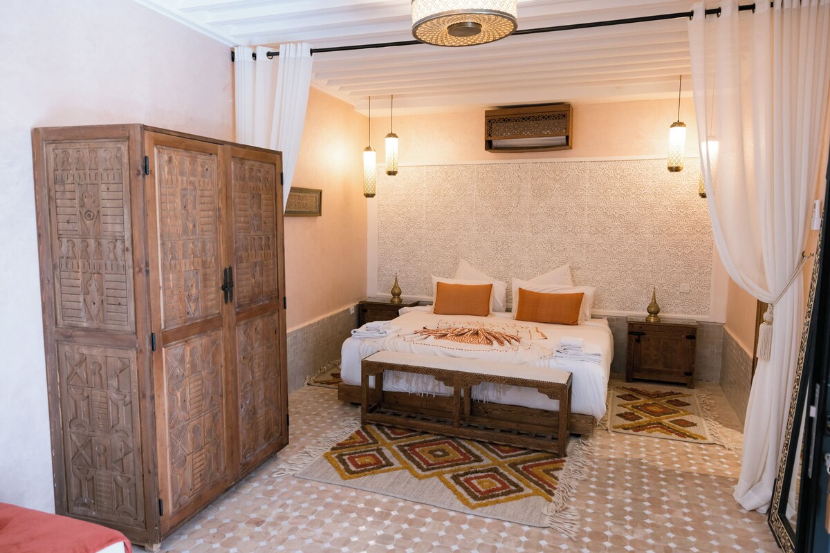A bedroom with a large wooden wardrobe, a bed with a patterned bedspread, two bedside tables, and hanging lamps.