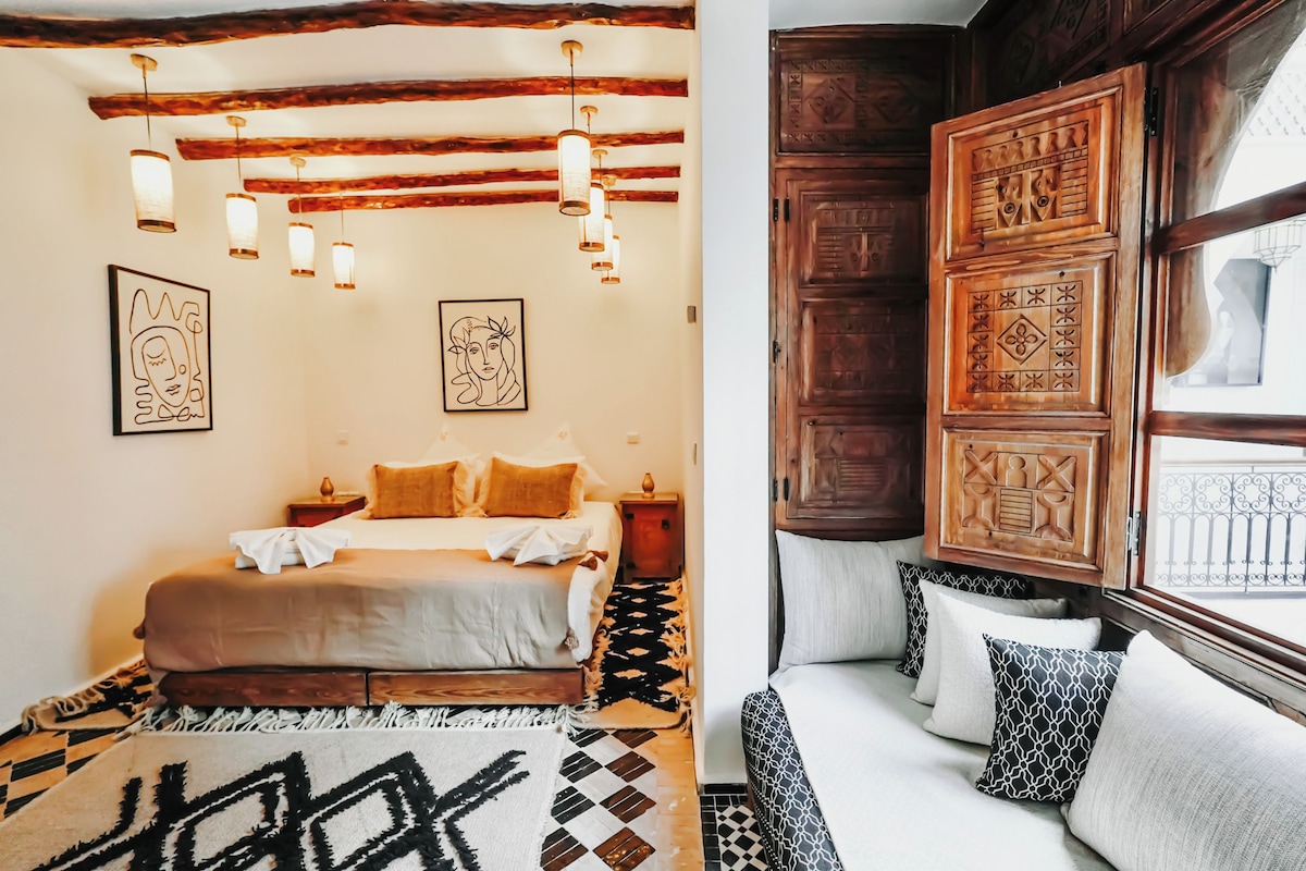 A bedroom with a bed, bedside tables, a patterned rug, and a window with decorative wooden panels.
