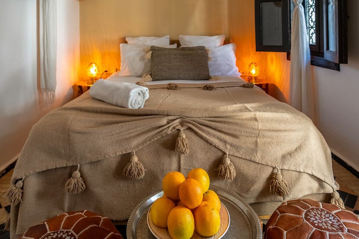 Una camera d'albergo con un letto grande, una lampada a soffitto decorativa, un elemento decorativo a forma di occhio sul muro e un vassoio con arance.