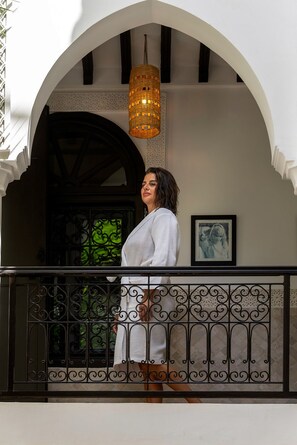A person standing on a balcony with a wrought iron railing, a wicker light fixture, and a framed picture on the wall.