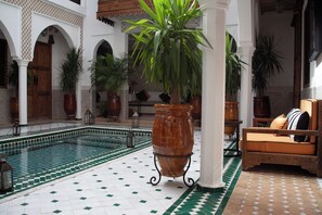 A pool area with a tiled floor, a large potted plant, and a sofa with cushions. - Riad Nelia Signature (Marrakech)