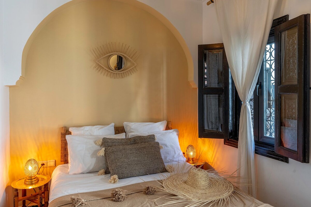 A bedroom with a large bed, a decorative eye motif on the wall, and a window with curtains.