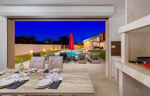 Un área de comedor al aire libre con vista a la piscina, sillas de descanso y un paraguas rojo.