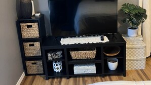 Une pièce avec un téléviseur posé sur un meuble noir, des paniers en osier, un élément décoratif mural et une plante en pot.