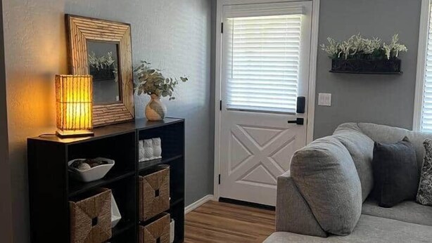 Une pièce avec un canapé gris, une console noire ornée de paniers, un lampadaire allumé et une porte blanche décorée d’une croix.