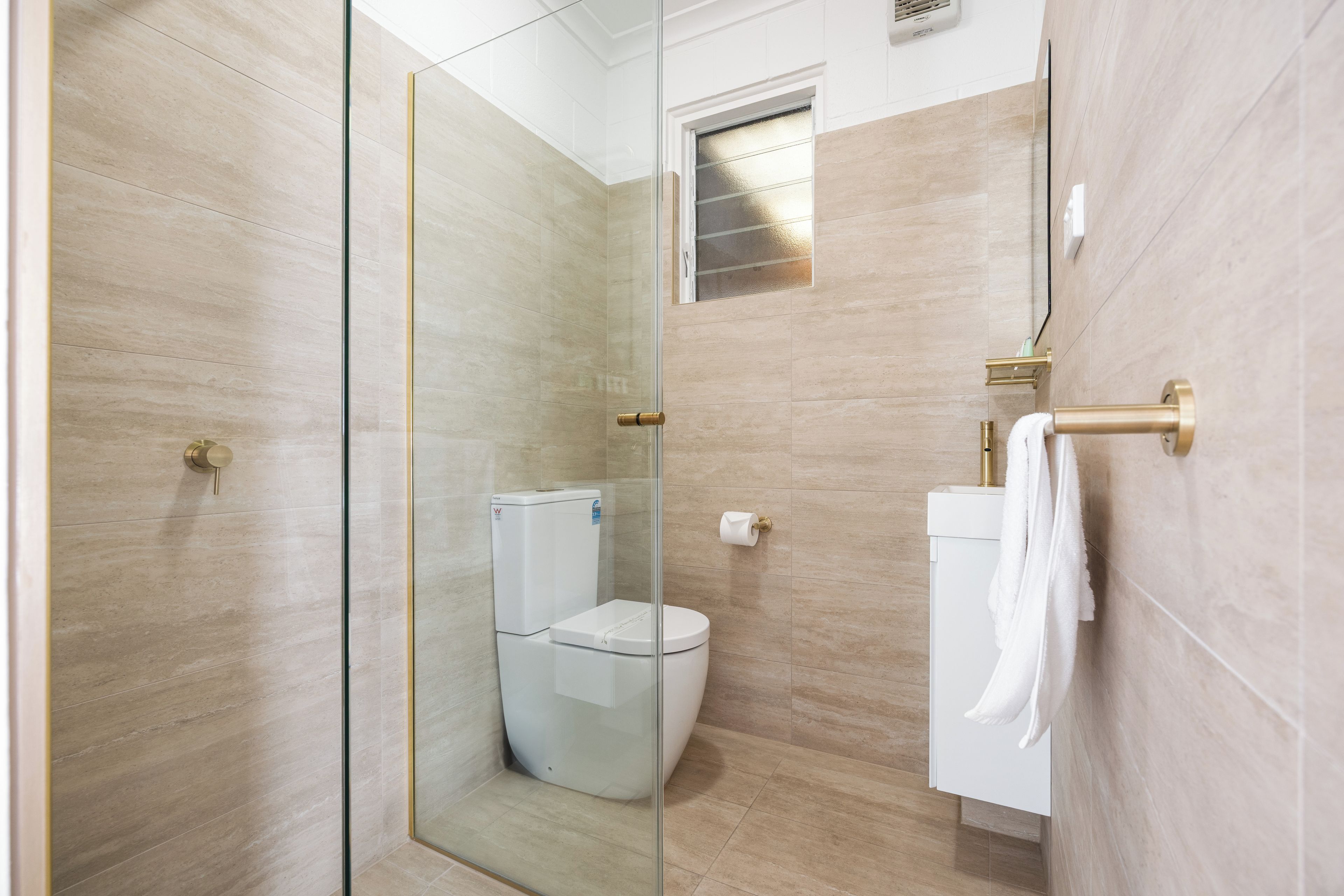Un baño moderno con cabina de ducha de vidrio, inodoro y lavamanos blanco, con una toalla colgada en la pared.
