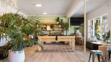 A pool table in a room with wooden flooring, surrounded by potted plants and shelves with books and decorative items.