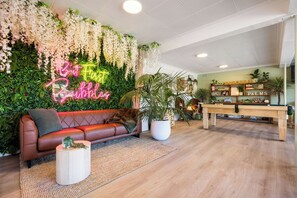 A modern lounge area with a leather sofa, a neon sign, a pool table, and lush greenery.