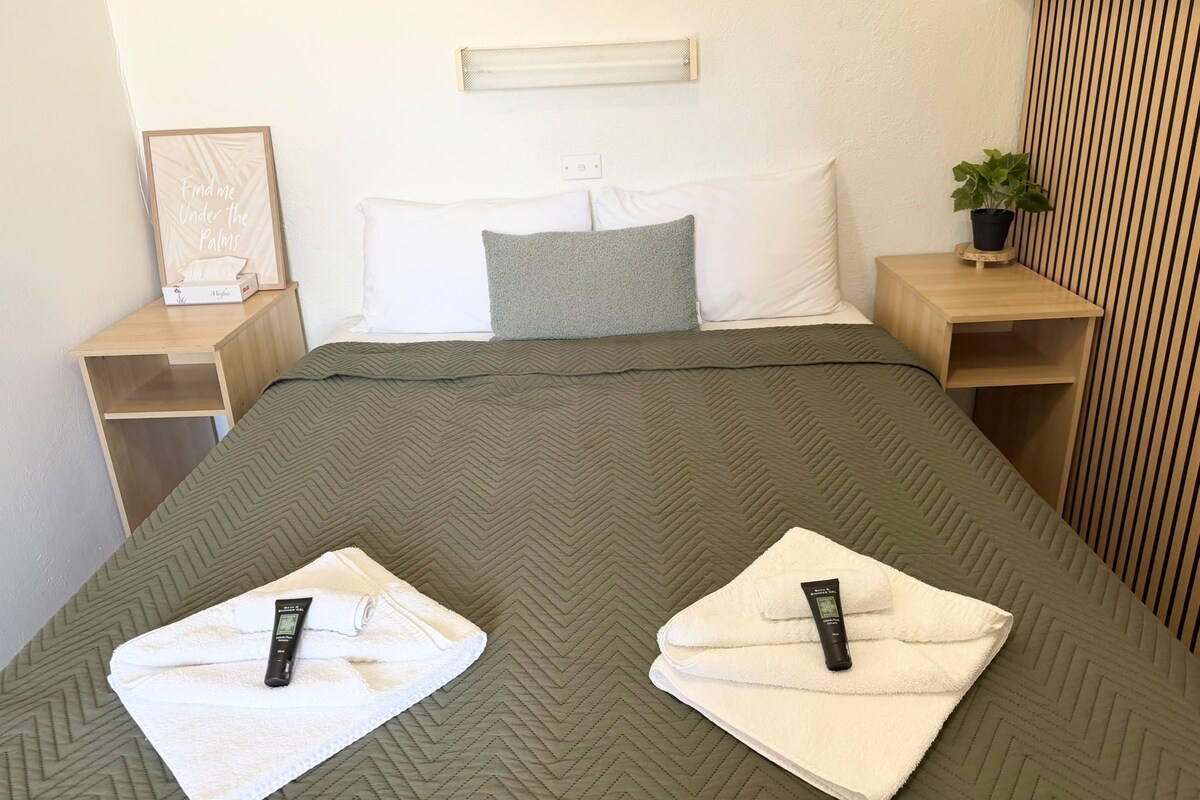 A hotel room with a bed, pillows, a bedside table, a nightstand, a wall-mounted light, and a potted plant.
