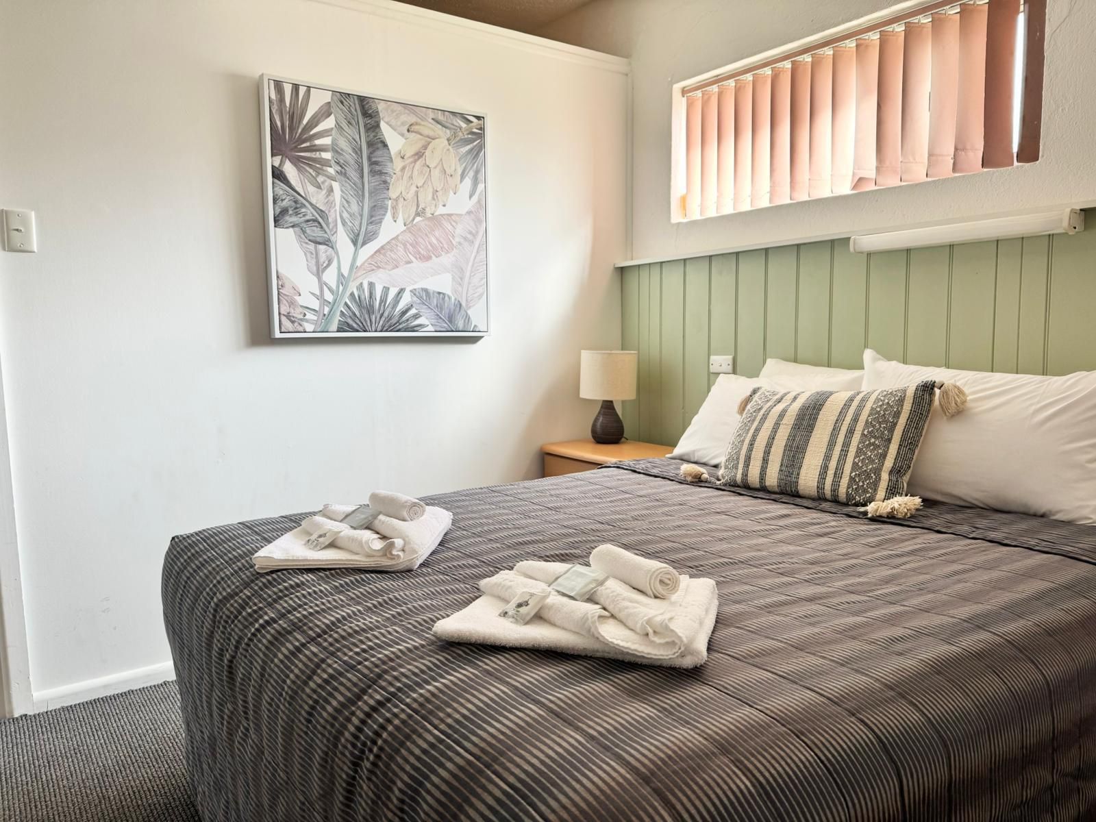 Un lit bien fait, avec une couvre-lit à rayures et des oreillers, une table de chevet avec une lampe, et un tableau aux motifs tropicaux accroché au mur.