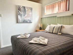 Un lit bien fait, avec une couvre-lit à rayures et des oreillers, une table de chevet avec une lampe, et un tableau aux motifs tropicaux accroché au mur.
