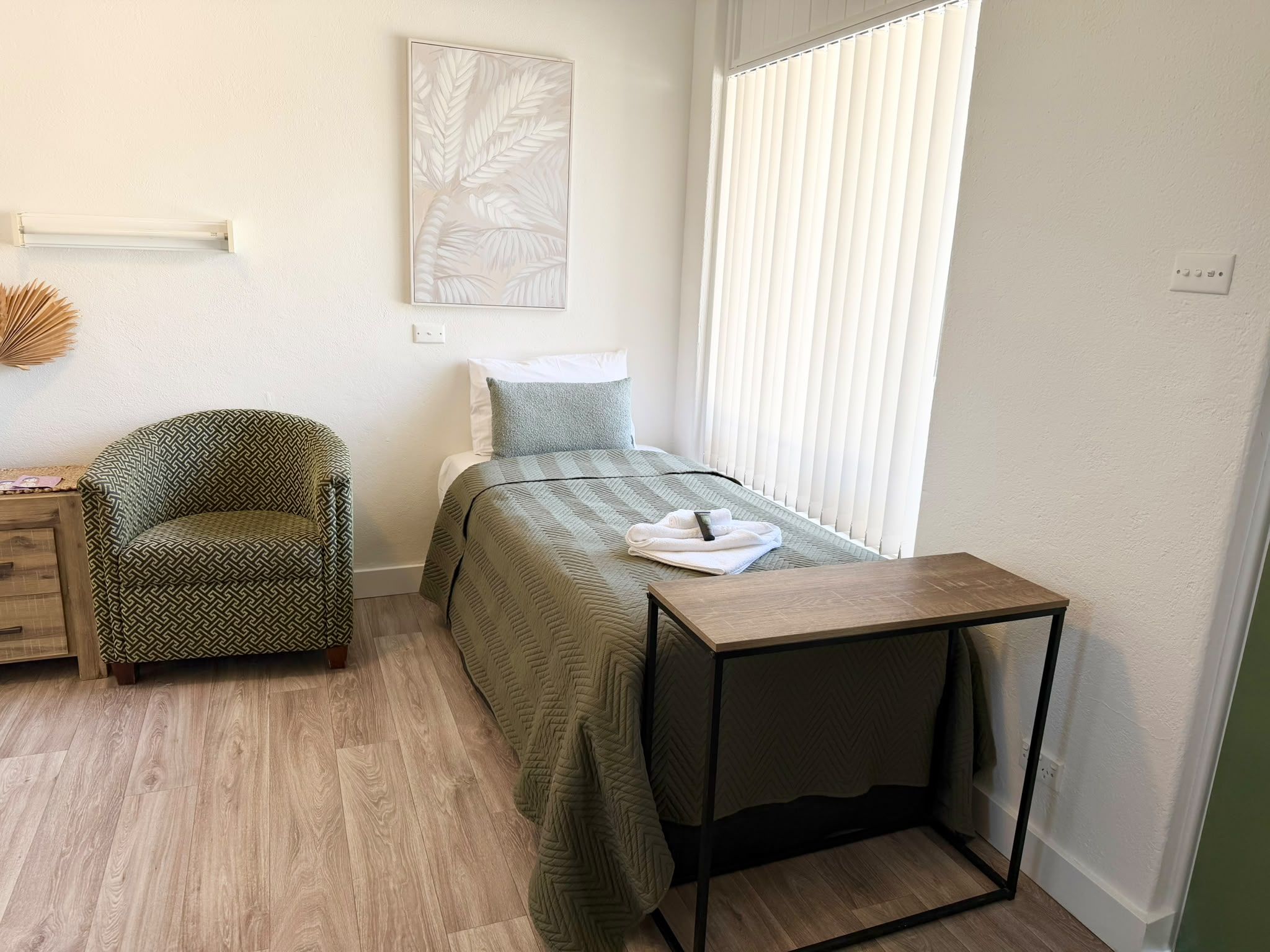 A small, neatly arranged bedroom with a bed, a chair, a bedside table, and a window with blinds.