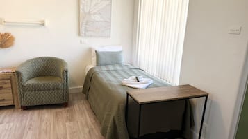 Ein kleines, ordentlich eingerichtetes Schlafzimmer mit Bett, Stuhl, Nachttisch und Fenster mit Jalousien.
