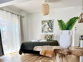 Un dormitorio con una cama, un ventanal con cortinas, una lámpara colgante, una obra de arte en la pared y una mesa de madera con una planta.