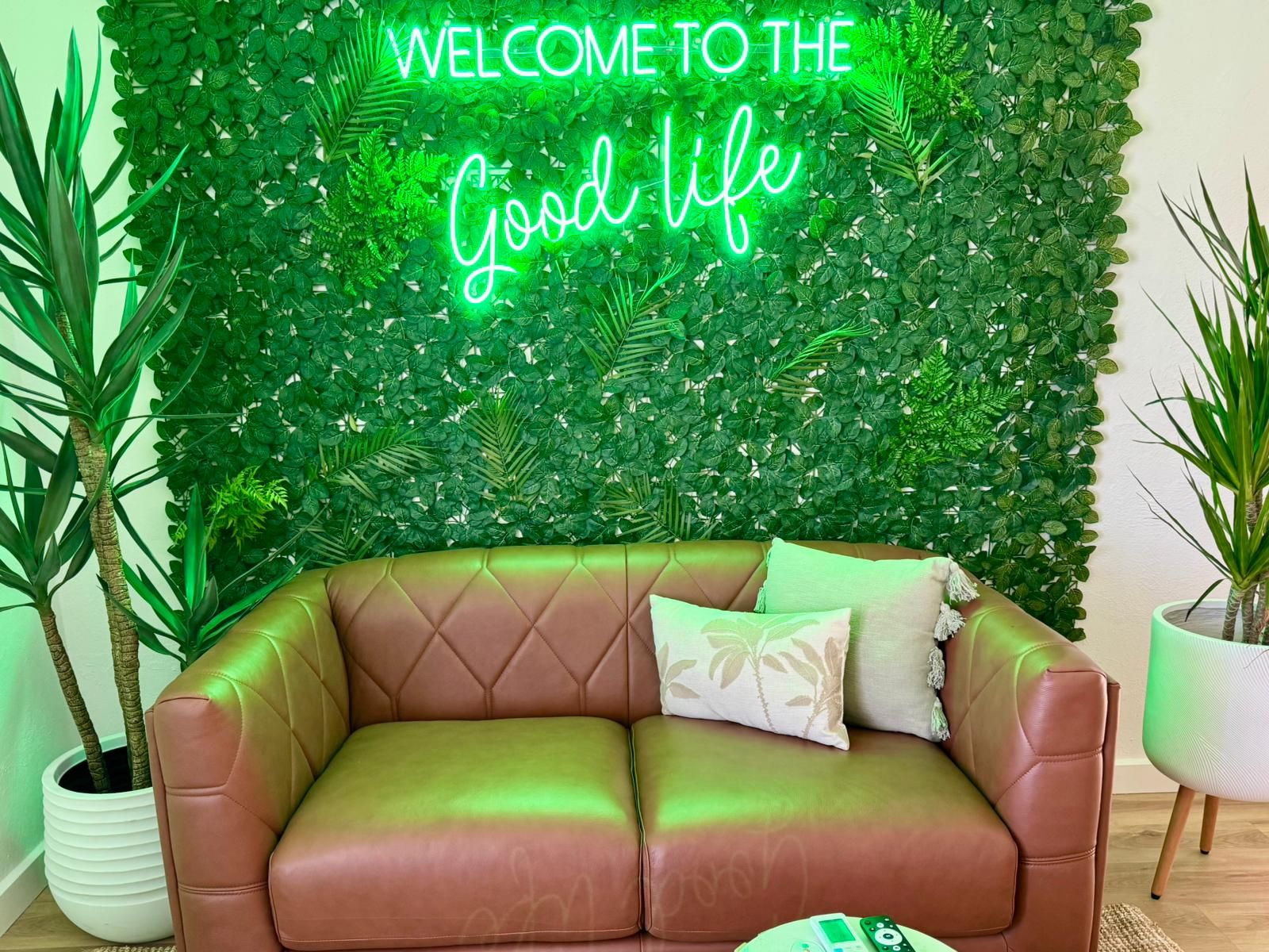 A brown leather sofa with a neon sign reading "WELCOME TO THE Good life" against a backdrop of green foliage.