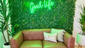 A brown leather sofa with a neon sign reading "WELCOME TO THE Good life" against a backdrop of green foliage.