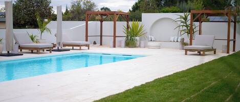 A swimming pool with lounge chairs and a pergola, surrounded by a white wall and greenery.