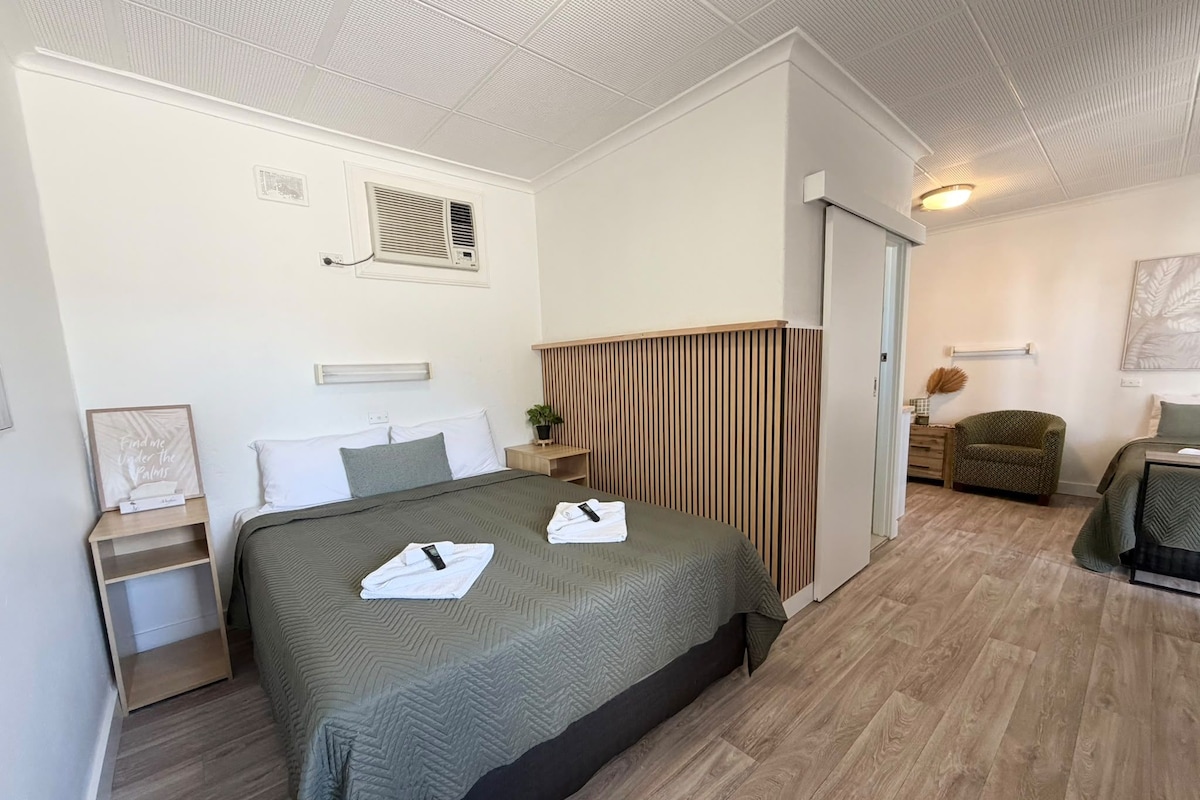 A hotel room with a bed, bedside tables, a plant, a chair, and a door leading to another room.