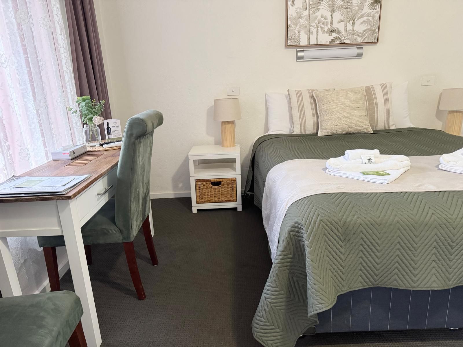 A hotel room with a bed, a desk, a chair, a nightstand, a lamp, and a wall-mounted artwork.